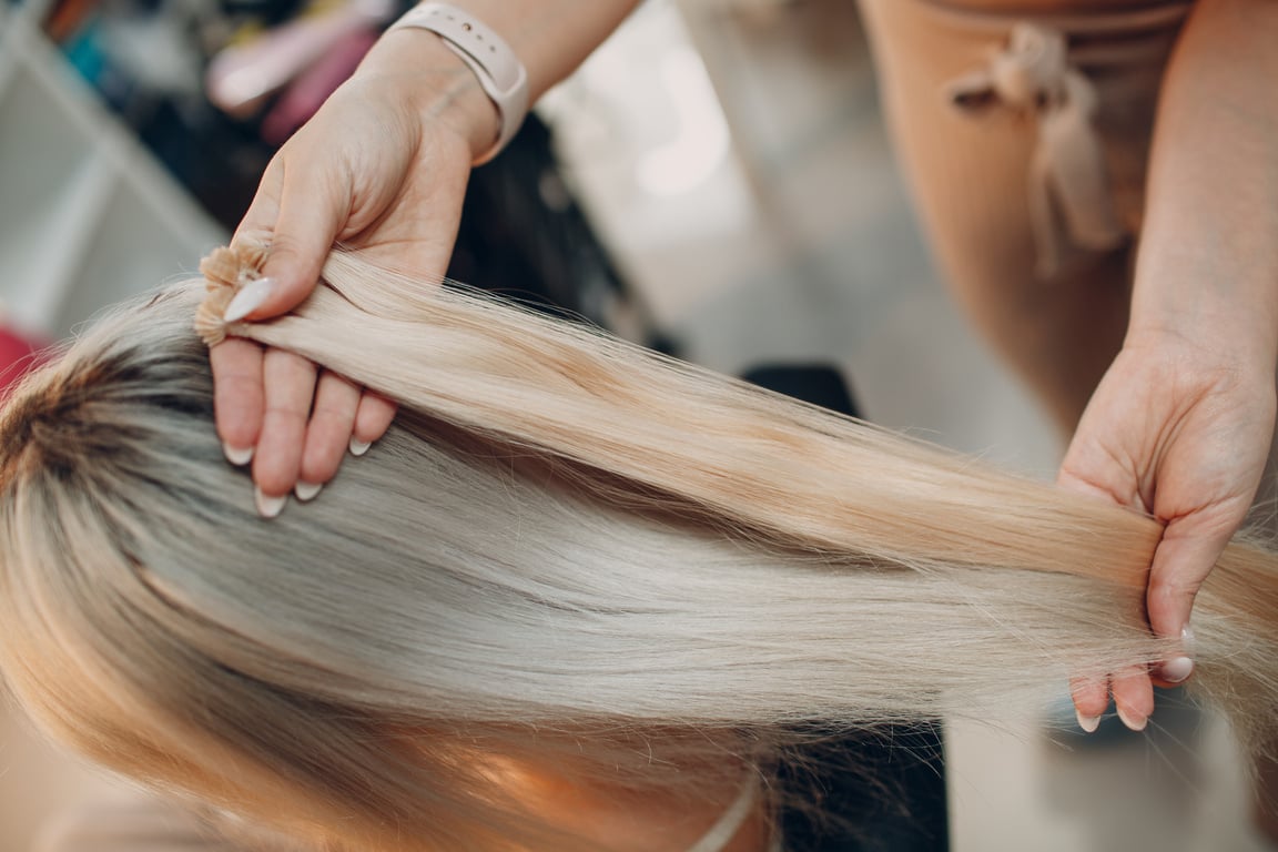 Hairdresser female making hair extensions to young woman with blonde hair in beauty salon. Professional hair extension strand of hair.